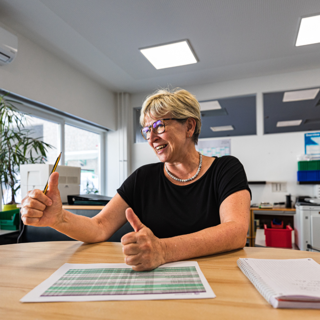 Fröhliche Frau mit blonden kurzen Haaren und Brille zeigt zwei Daumen hoch.