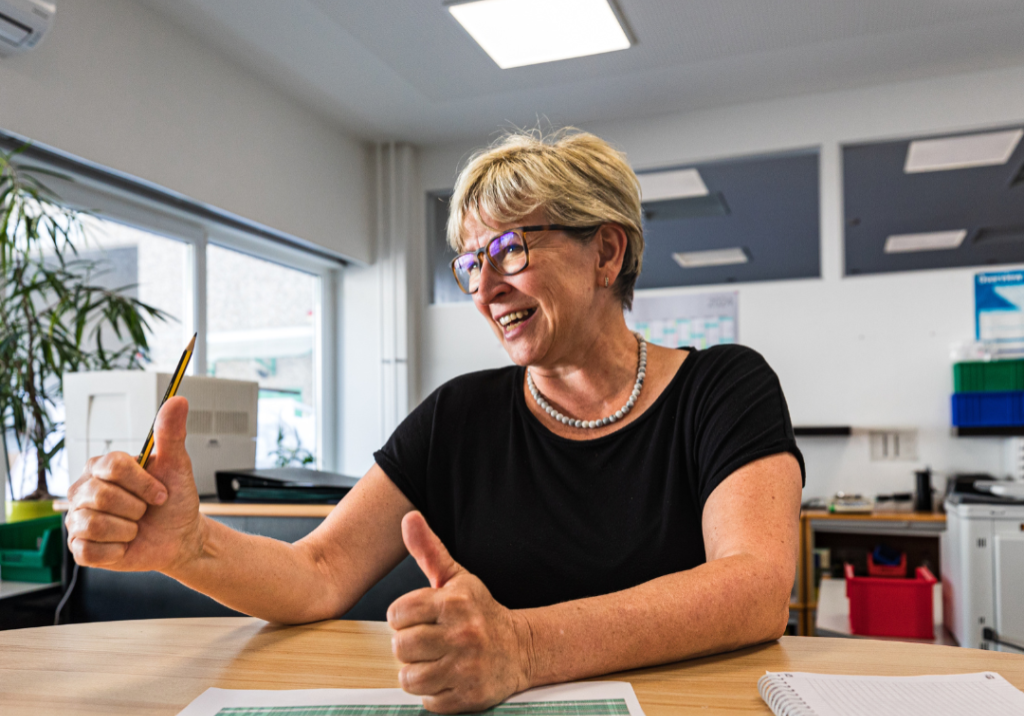 Fröhliche Frau mit blonden kurzen Haaren und Brille zeigt zwei Daumen hoch.